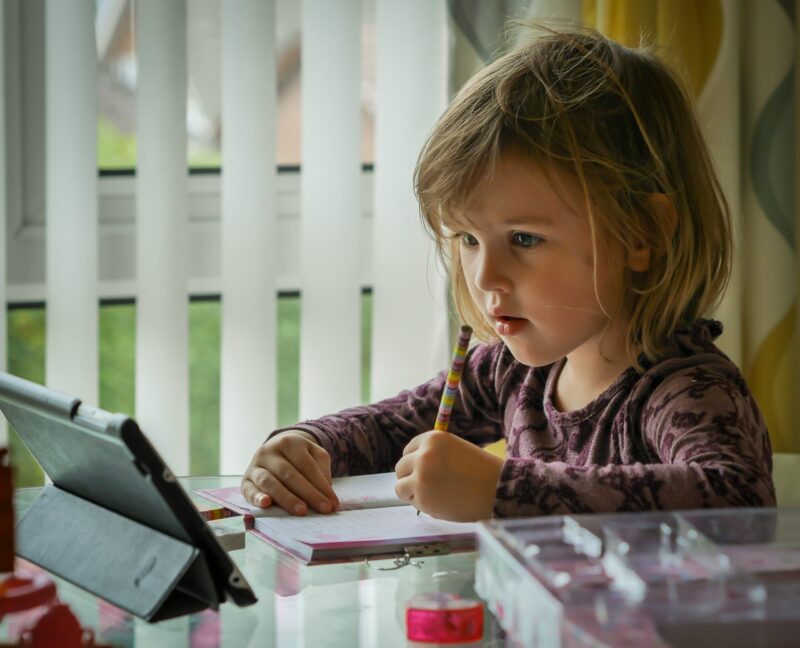 Young child going school work, working in front of a tablet computer with a pencil and paper at the ready.