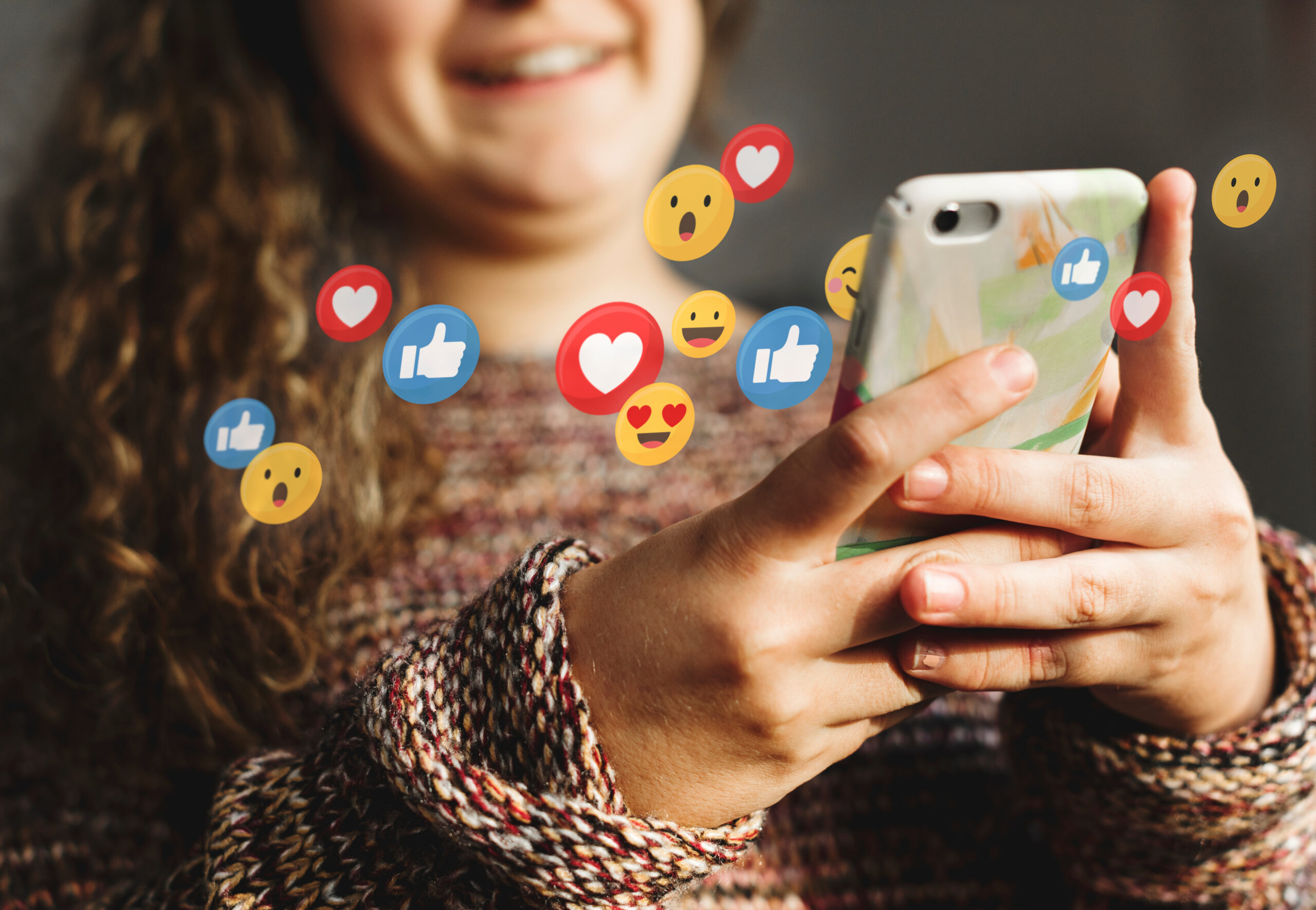 A young girl holding and looking at her phone as social media reactions spill forth from it.