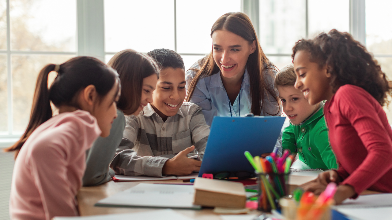 A teacher leads a class gathered around from a laptop on the table in front of them