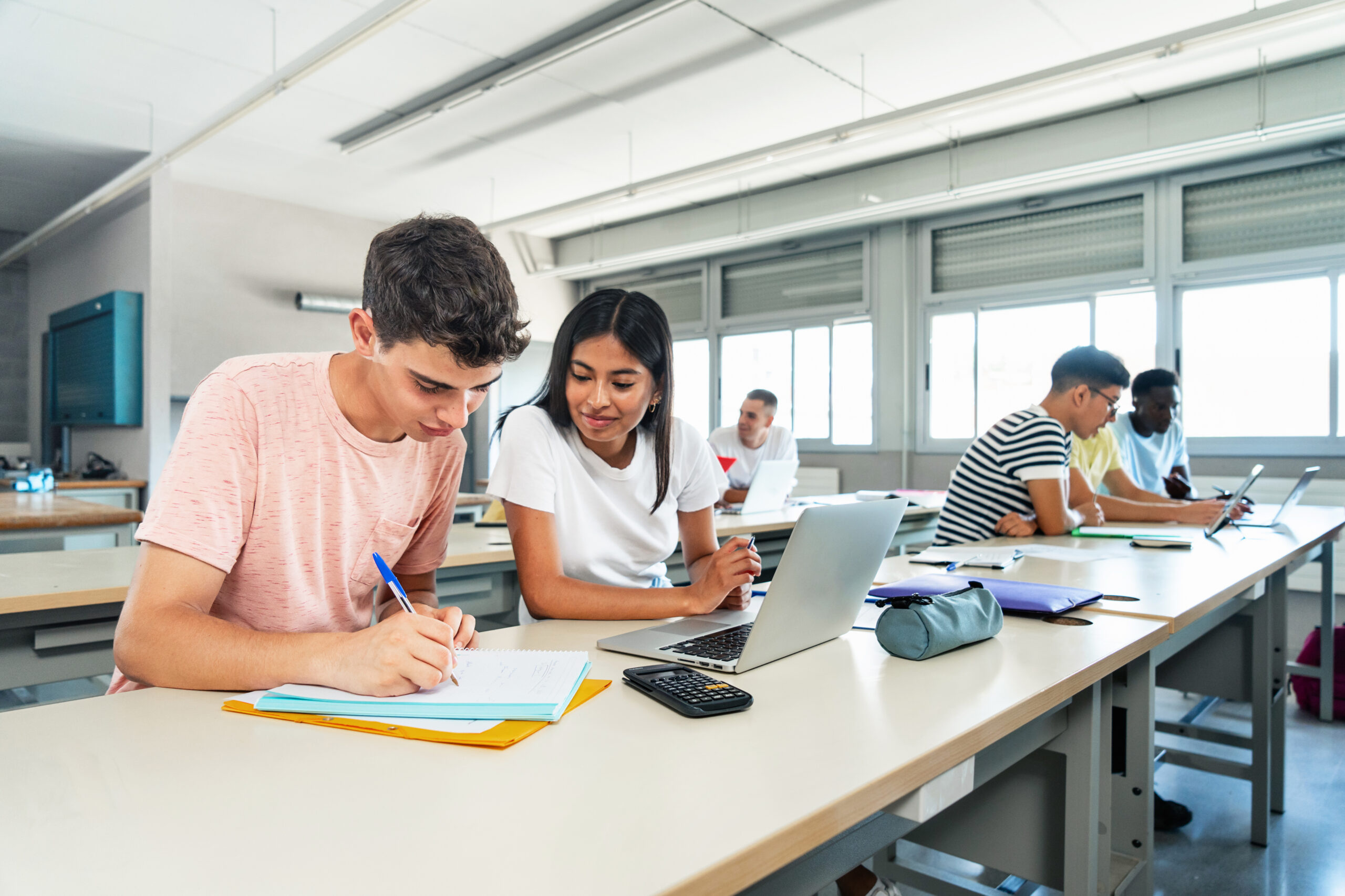 students at a class table, most using laptops but one using pen and paper